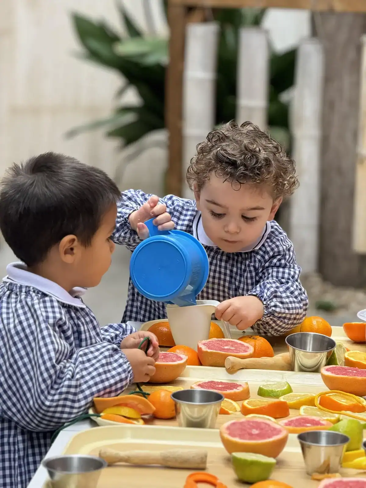 Two young children pouring liquid into bowls at a table set with fruits.