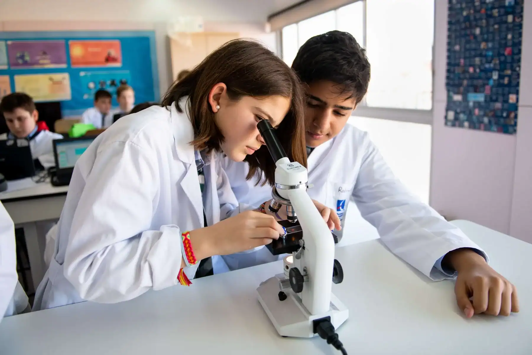 Two students in lab coats looking through a microscope in a science classroom.