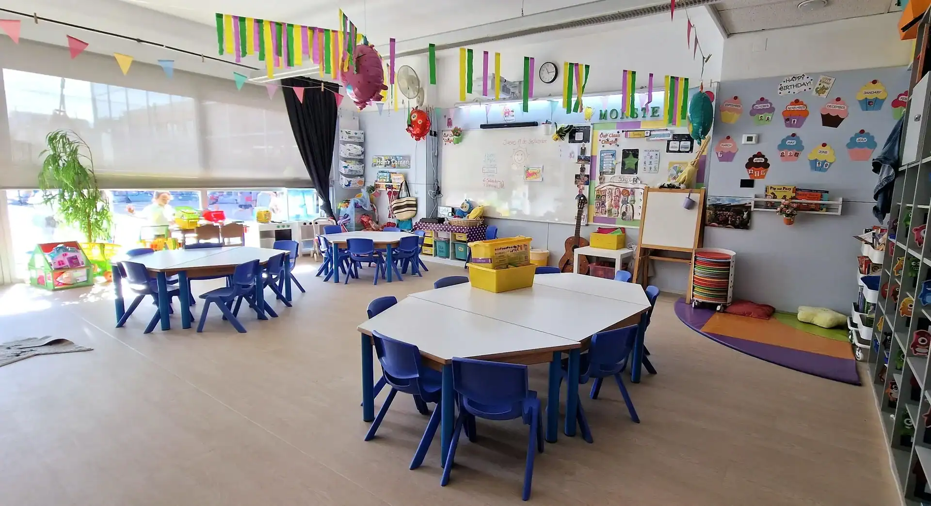 Bright classroom with colorful tables and chairs for children, decorations, and play area.