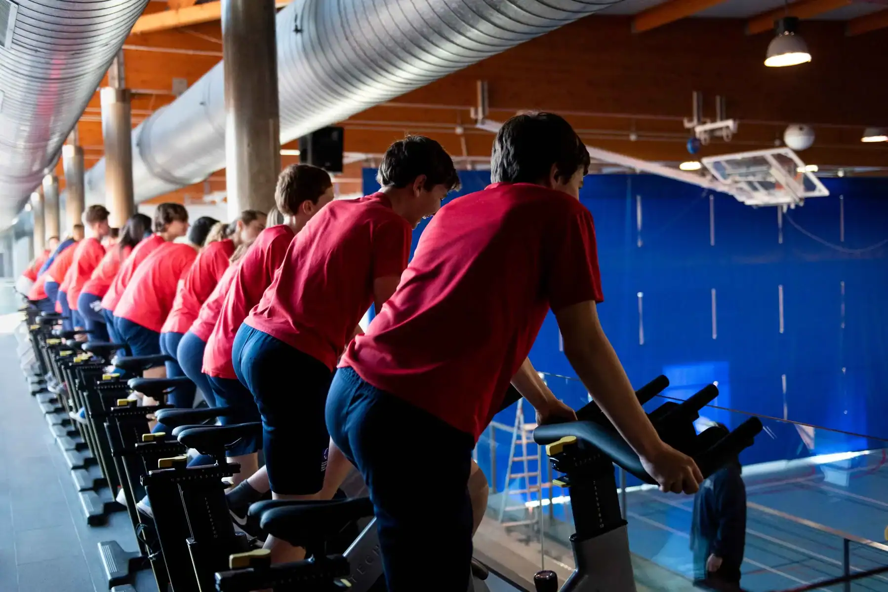 Group of students in red shirts cycling on stationary bikes indoors.