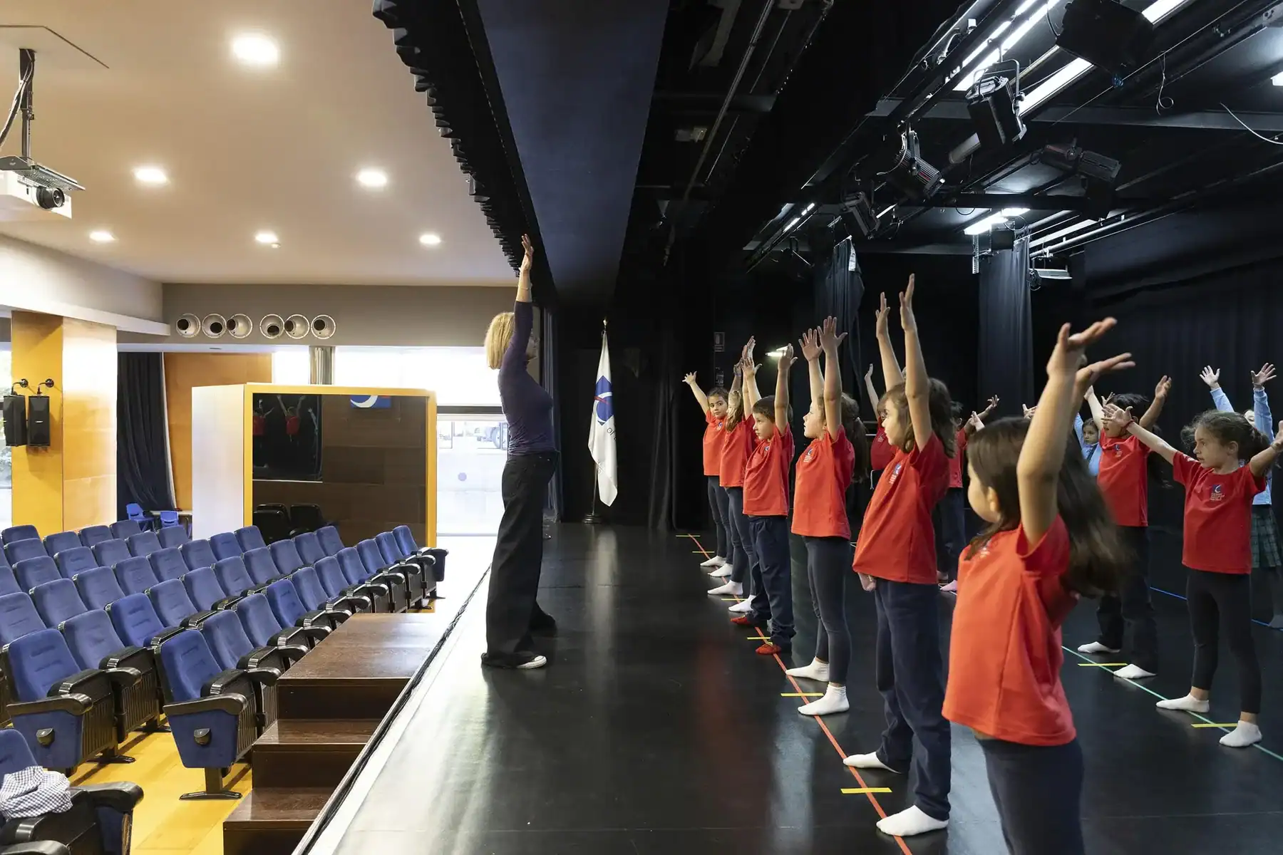 Students in red shirts raise arms during a stage rehearsal at a theater.