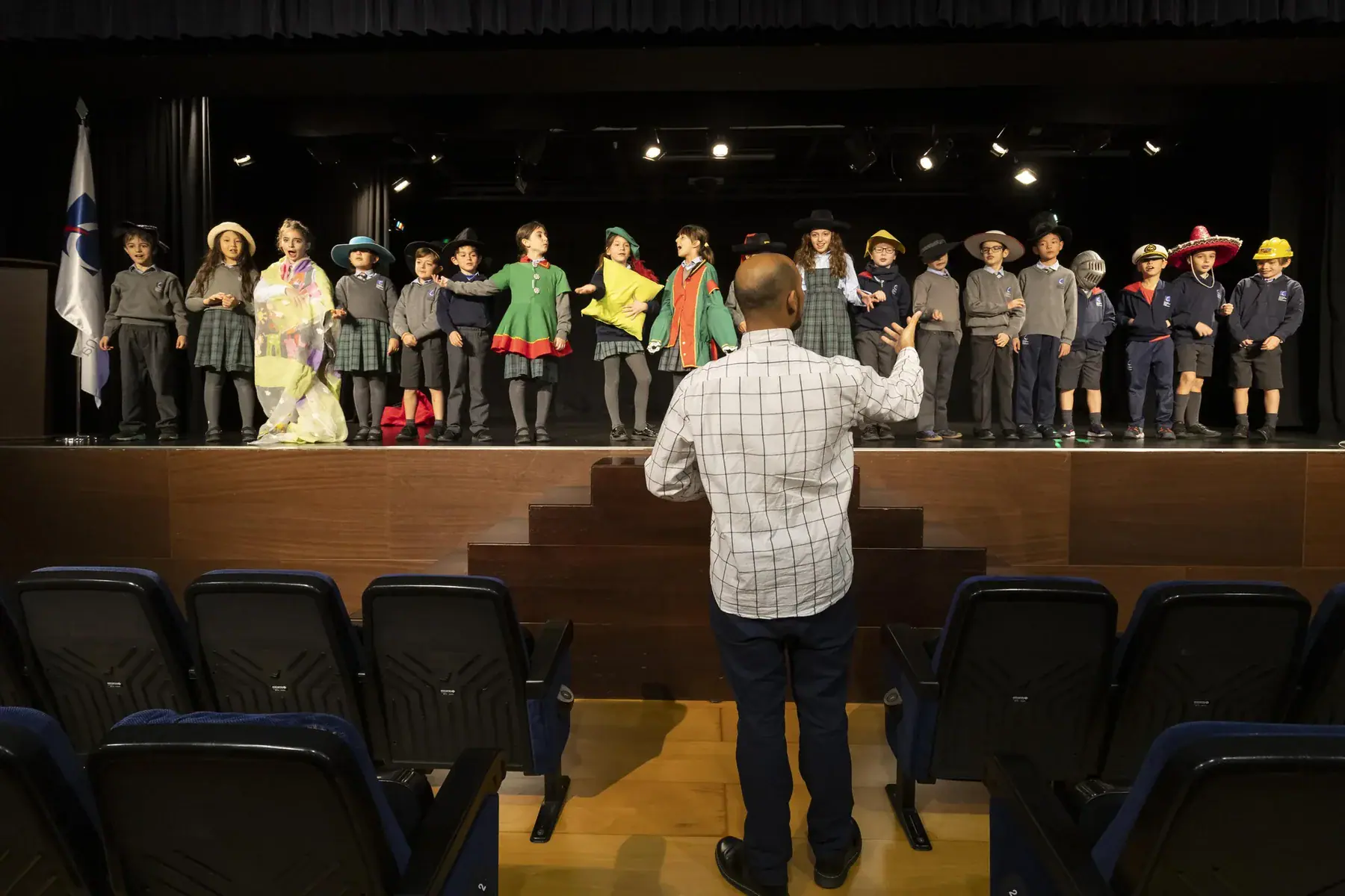 Group of children on a stage rehearsing with a conductor in a theater.