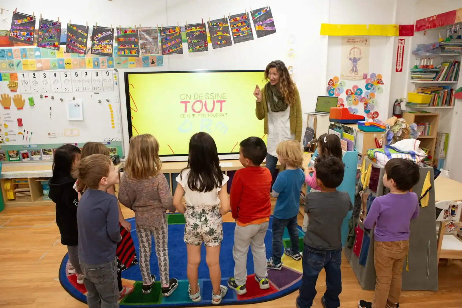 Young students gathered around a teacher at a colorful classroom screen.