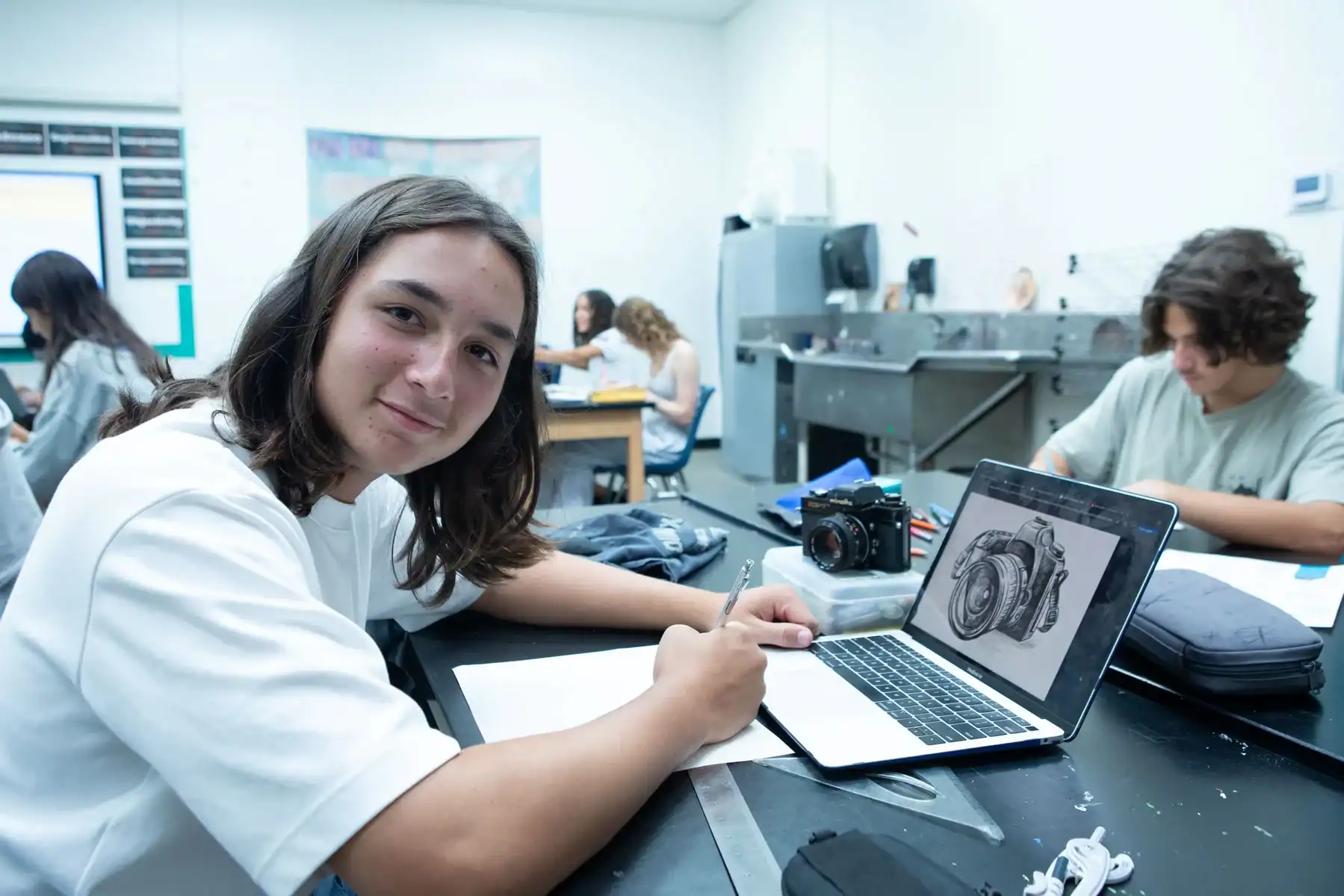 Student drawing on laptop in a classroom with photography equipment.