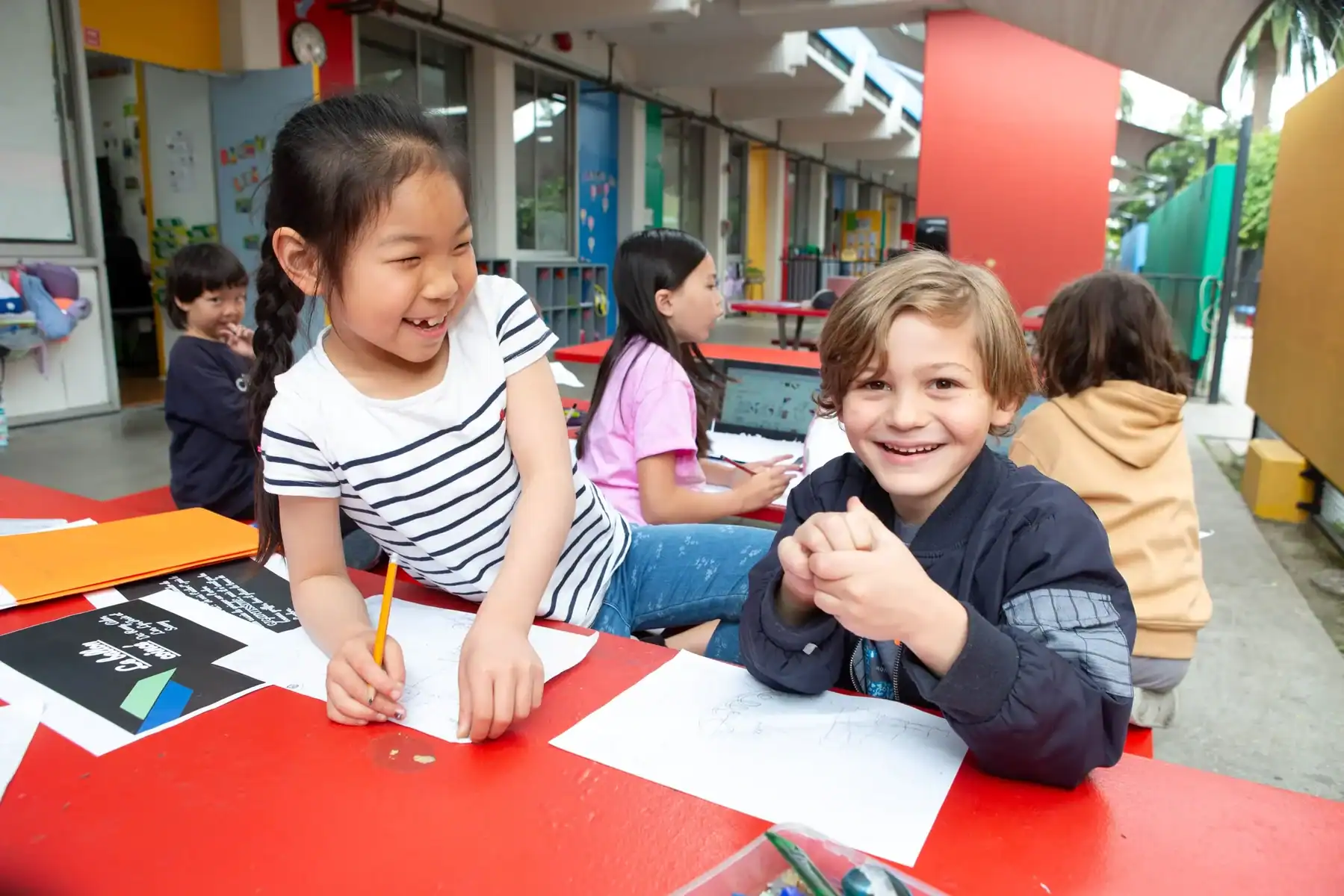 Two smiling children drawing at a colorful outdoor classroom with pencils and paper.