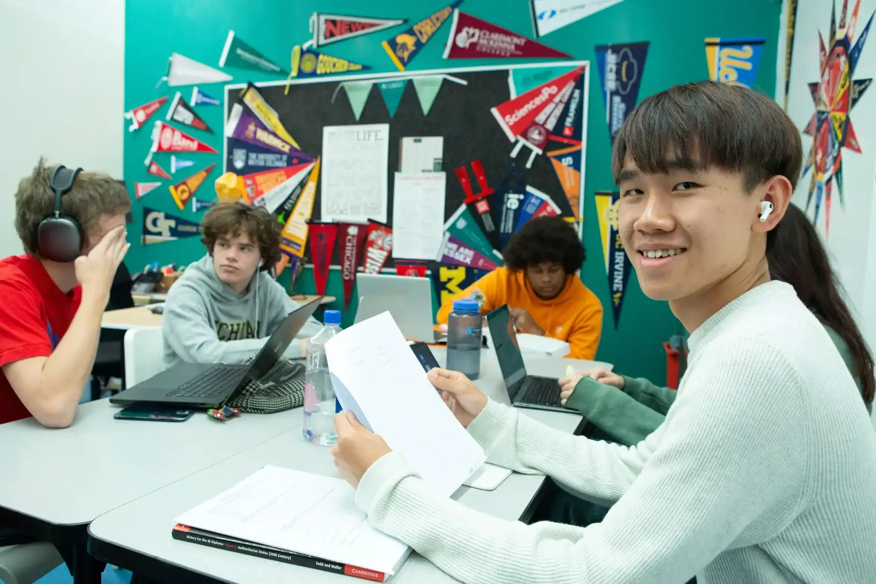 Students in a classroom collaborate with laptops and papers, smiling.