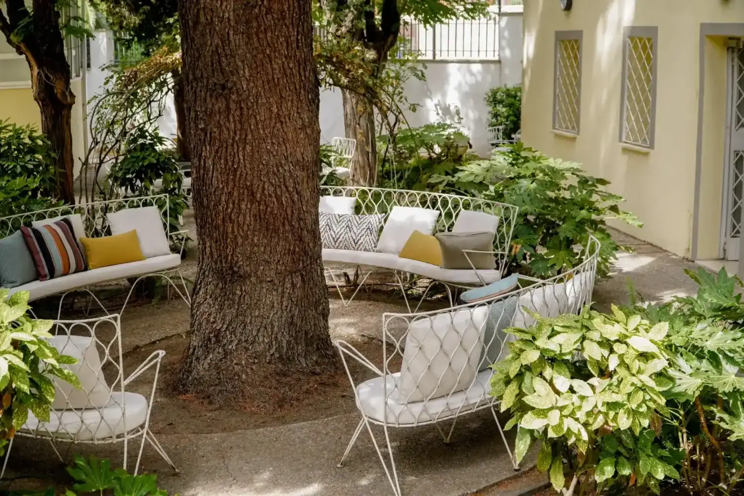 Outdoor garden seating with white metal chairs and colorful cushions under a large tree.