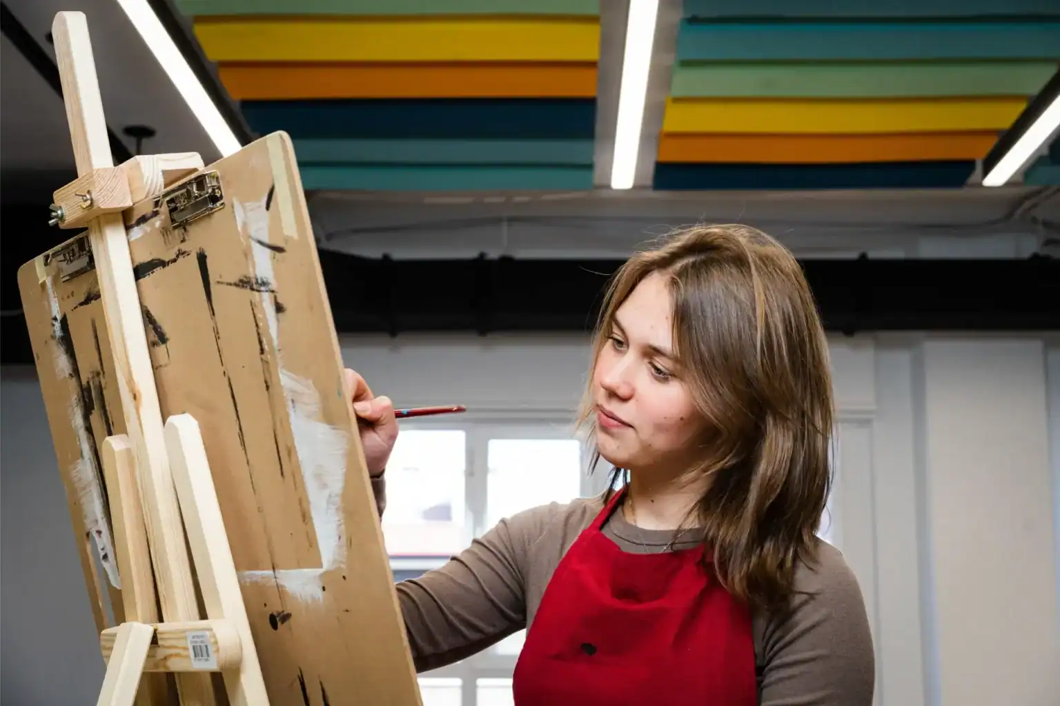 Young woman artist working on a painting at an easel in a modern classroom setting.
