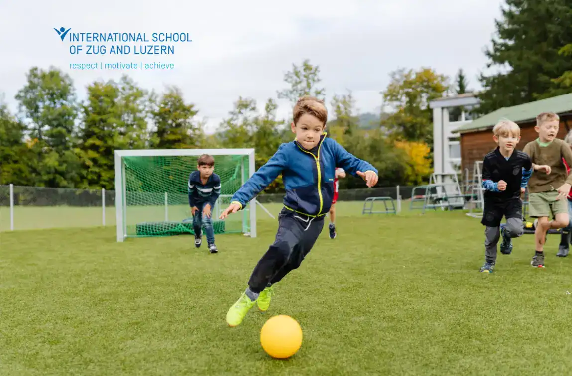 Un groupe d'enfants joue au football sur un terrain vert. Un garçon au premier plan court énergiquement vers un ballon jaune, tandis que d'autres le poursuivent. Le logo de l'École internationale de Zoug et de Lucerne est visible dans le coin supérieur gauche.