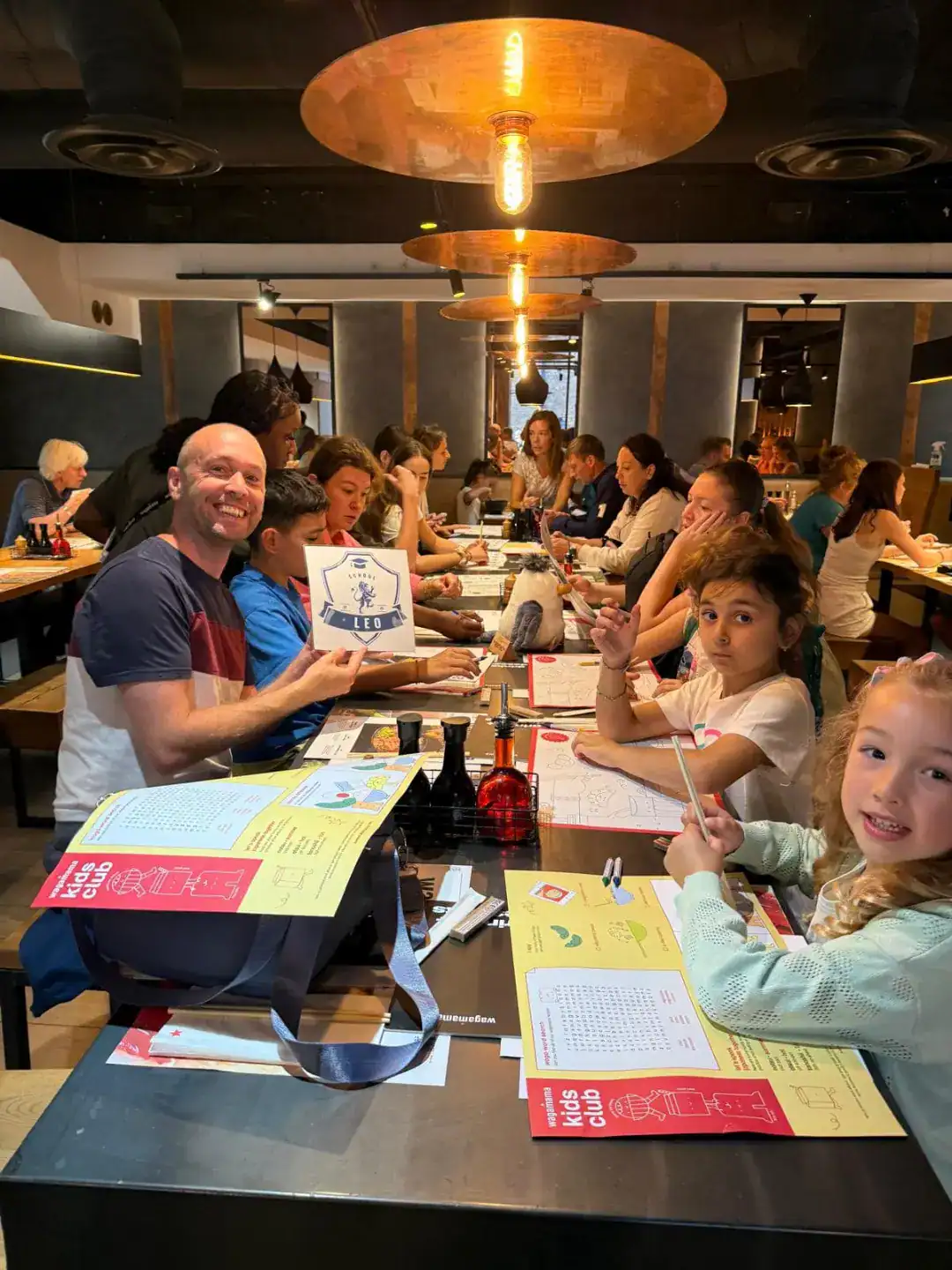 Bright classroom scene at a World Schools event with students engaging in activities and a cheerful instructor holding a Kids Club sign.