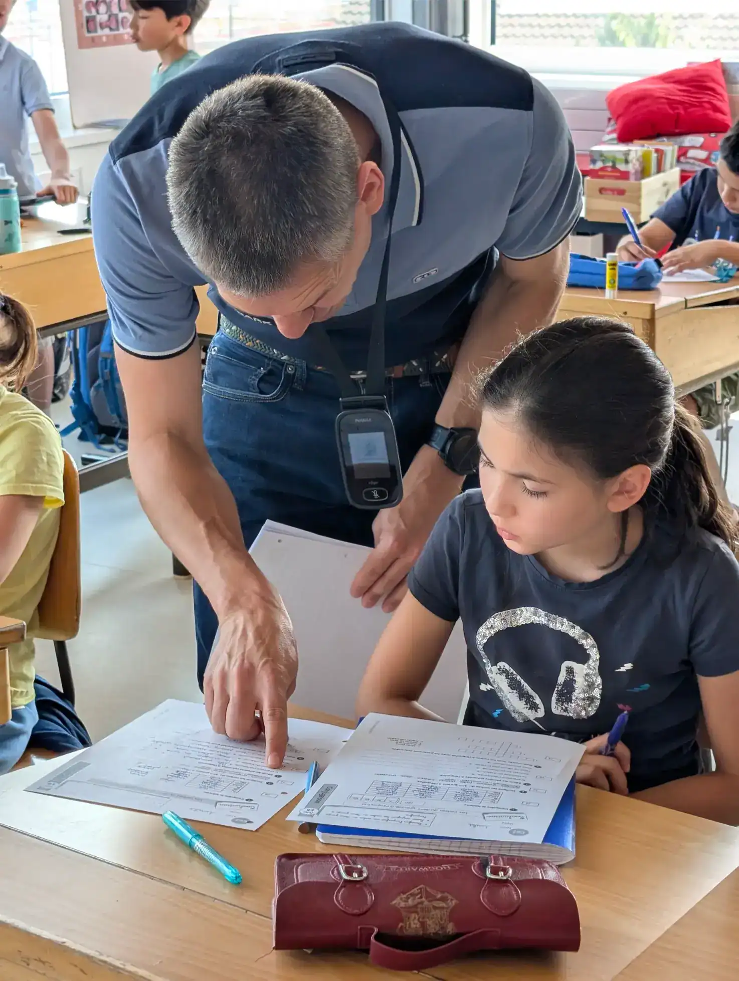 Focused teacher assisting a student with schoolwork in a diverse classroom setting.