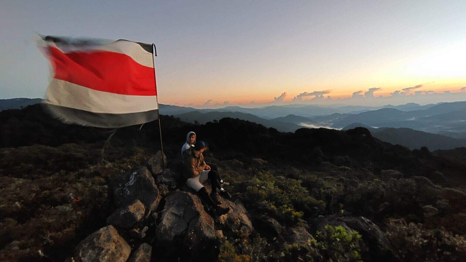 1. People sitting on rocks with a flag on a mountain at sunrise, scenic view of distant mountains.
