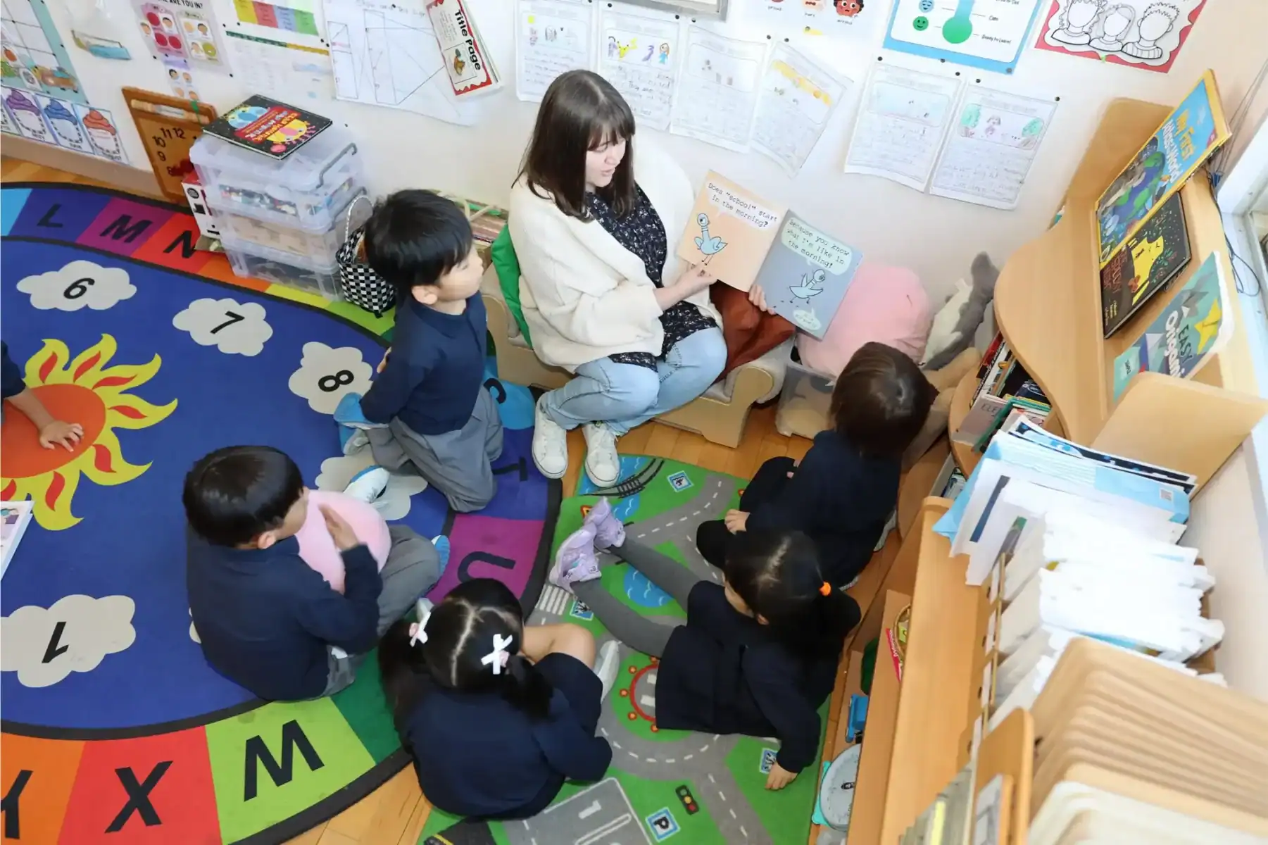 A teacher reads a children's book to a small group of students in a colorful classroom.