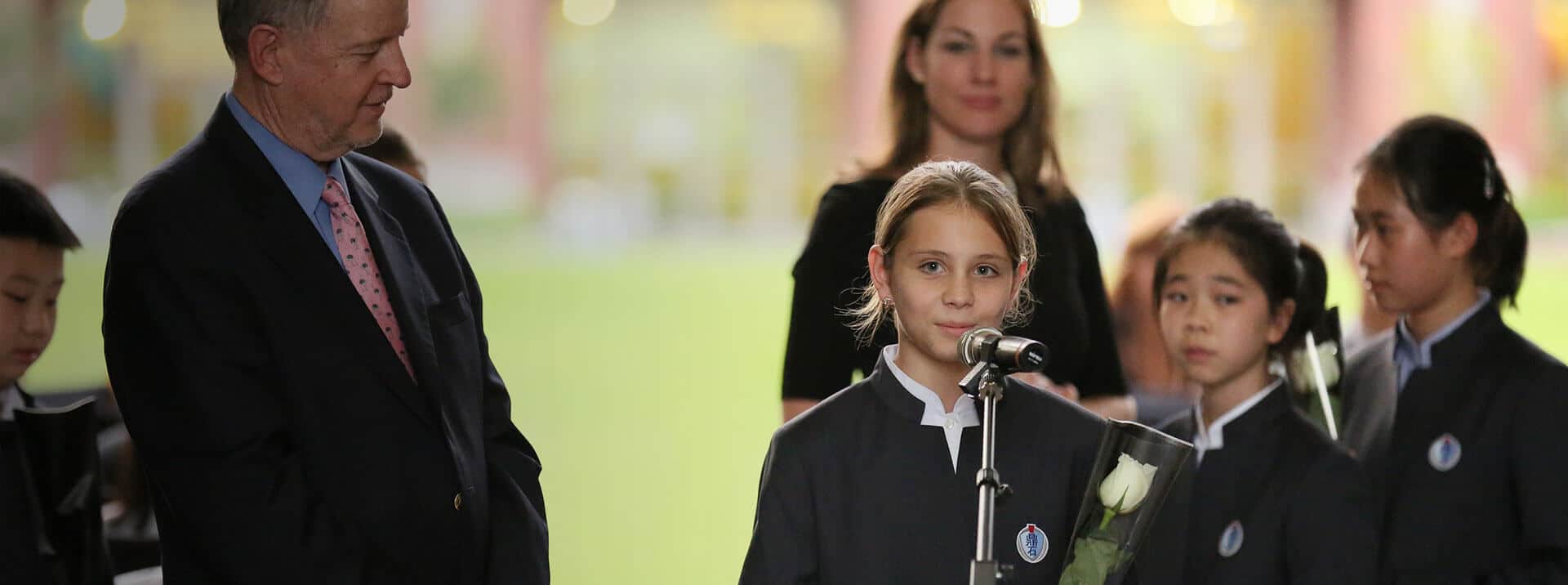Young girl speaking at school event, surrounded by students and teachers, celebrating academic achievement.