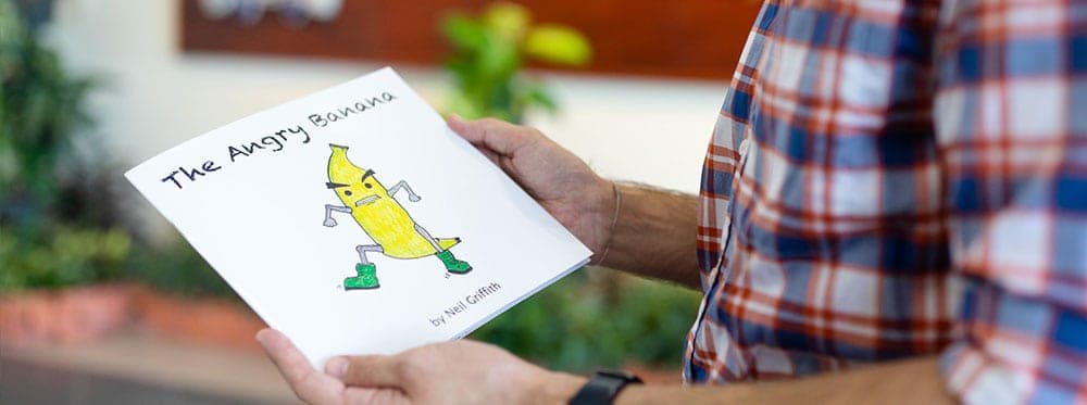 Happy person reading a children's book titled "The Angry Banana" outdoors in a school setting, promoting literacy.