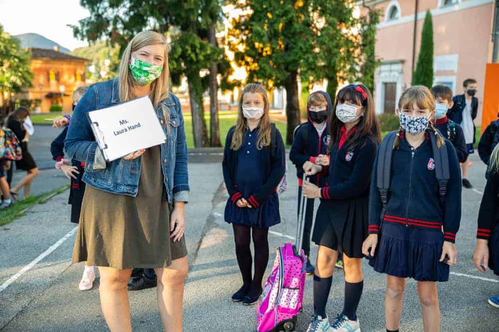 This Month at TASIS: September 2020 7 Young students with backpacks wearing masks, standing outdoors with a teacher in a school uniform.