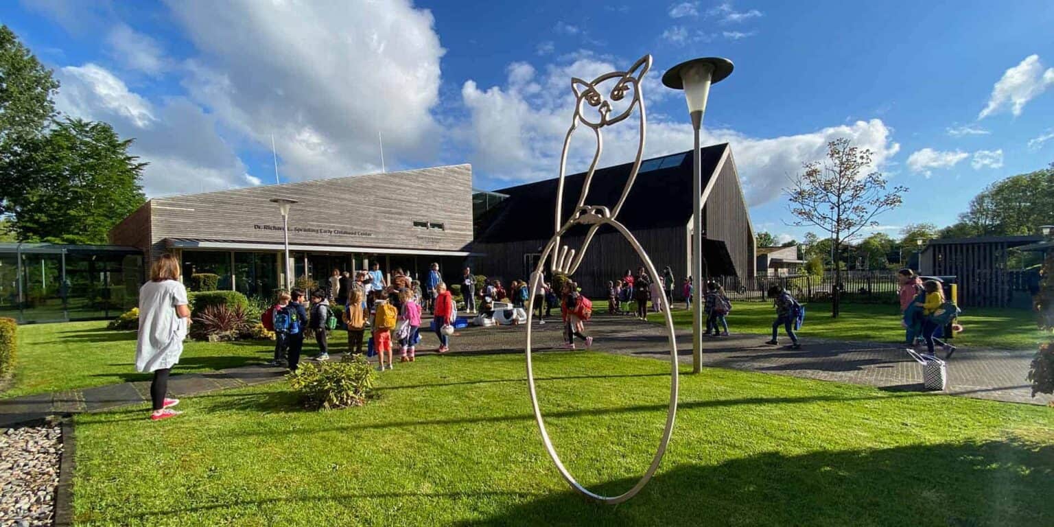 Brightly lit childhood education center with children arriving, vibrant greenery, and unique street art sculpture.