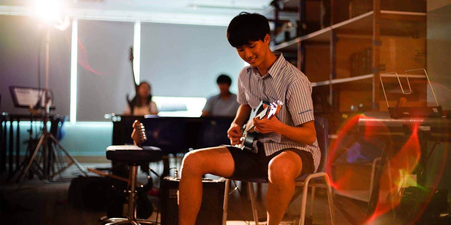 Boy playing electric guitar in a music classroom with students in the background, bright stage lighting.