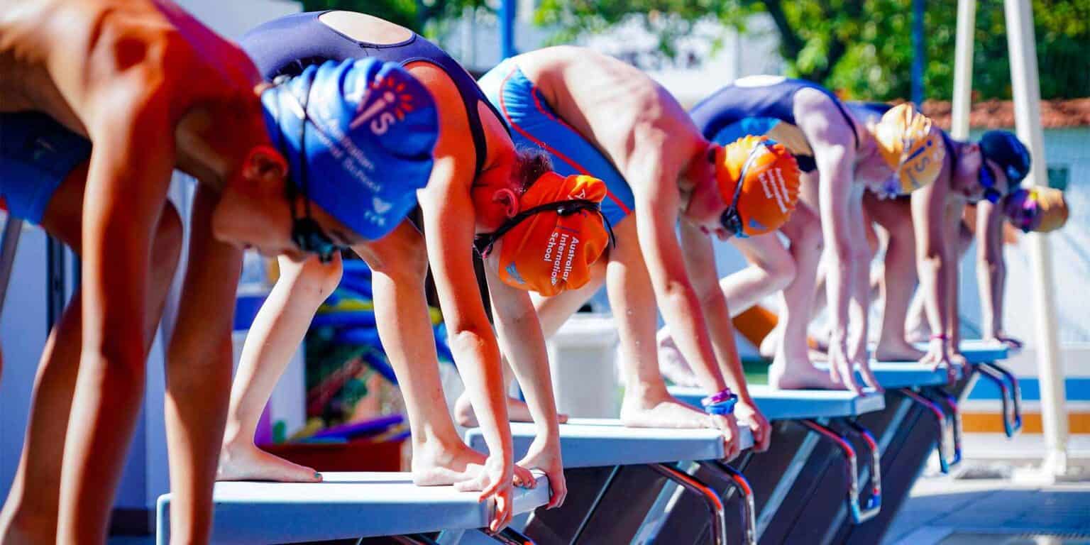 Children in colorful swimming caps and swimsuits prepare to race at a school swimming competition.