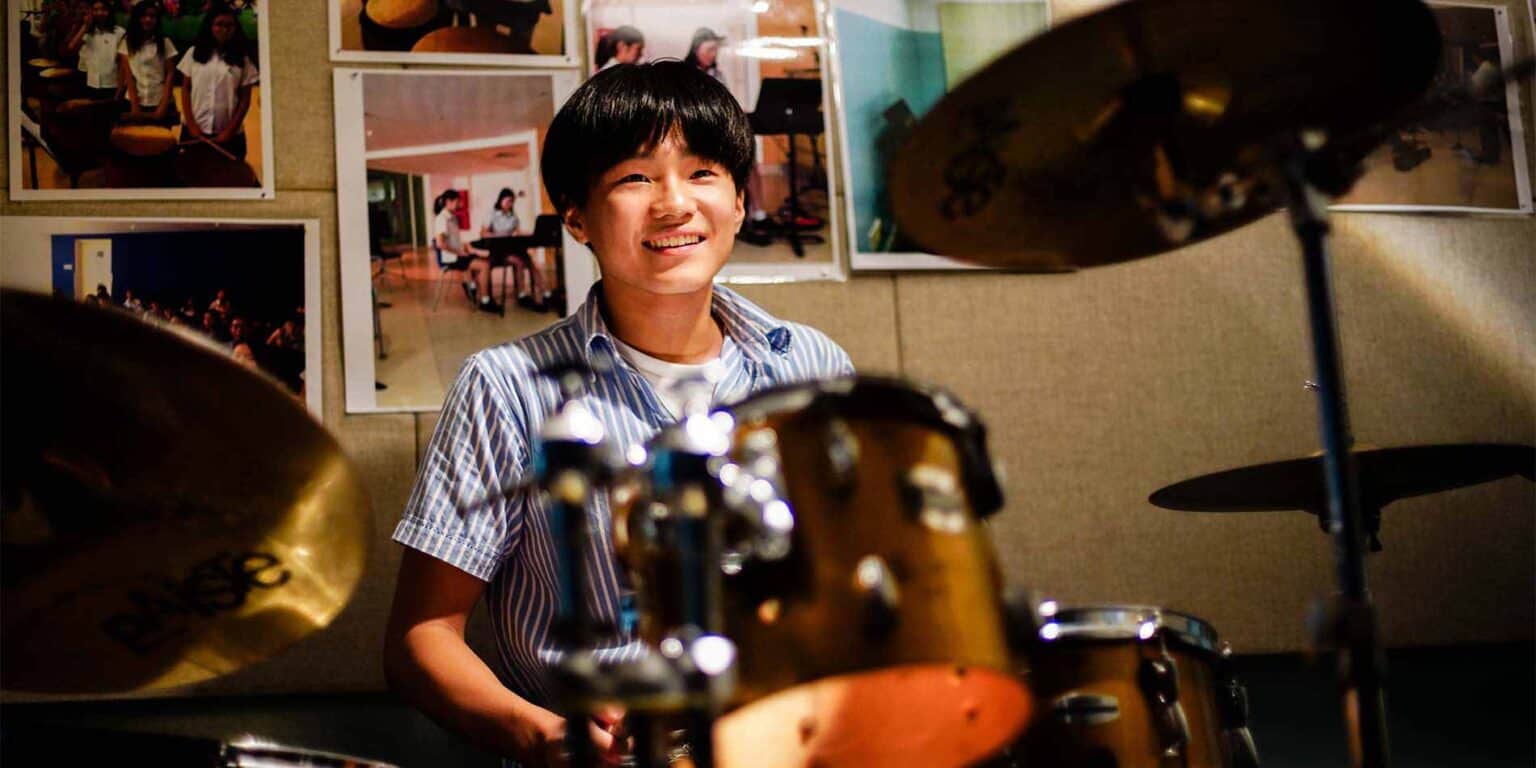 Young boy smiling behind a drum set in a music classroom at a world school.