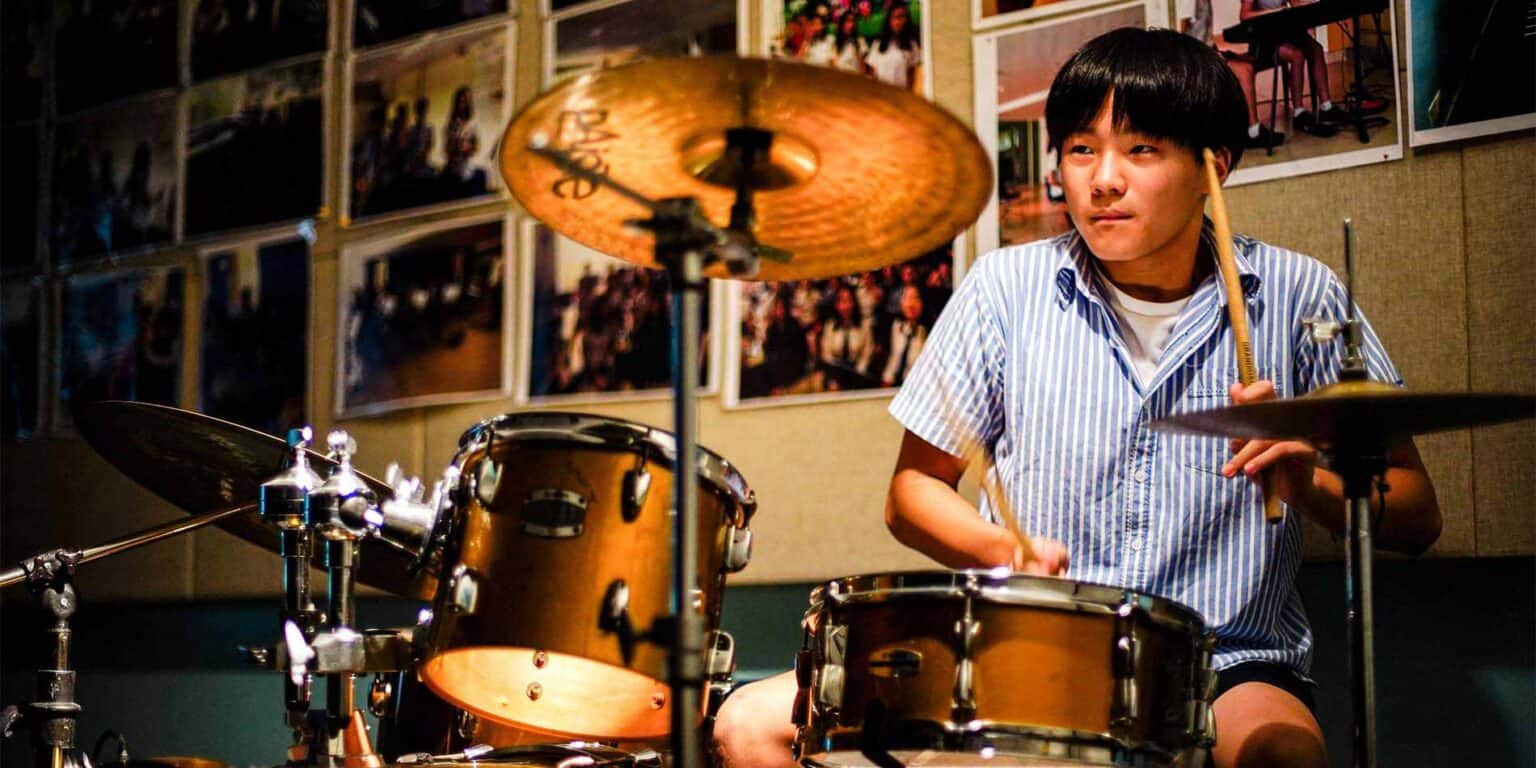 Young male student playing drum set in school music class, focus on drum kit and cymbals.