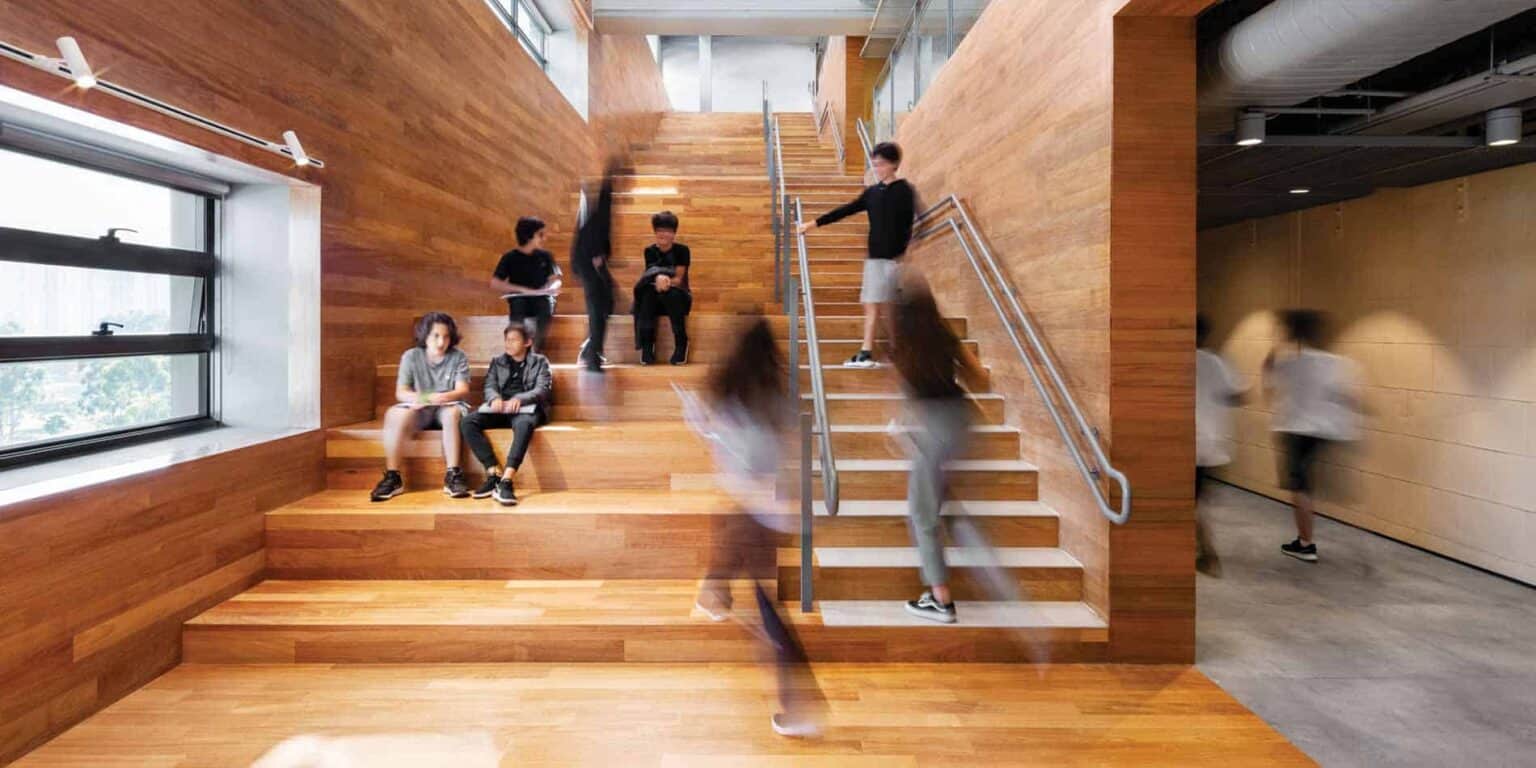 Bright modern school interior with students sitting and walking on wooden stairs, lighting and glass windows.