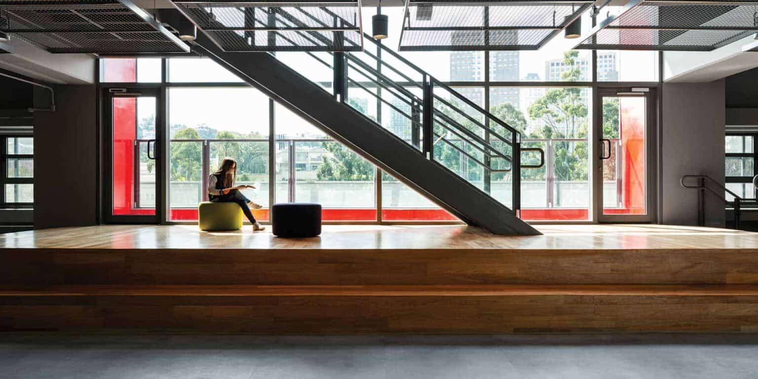 Bright modern school lobby with large windows, staircase, and student lounging on seating area.