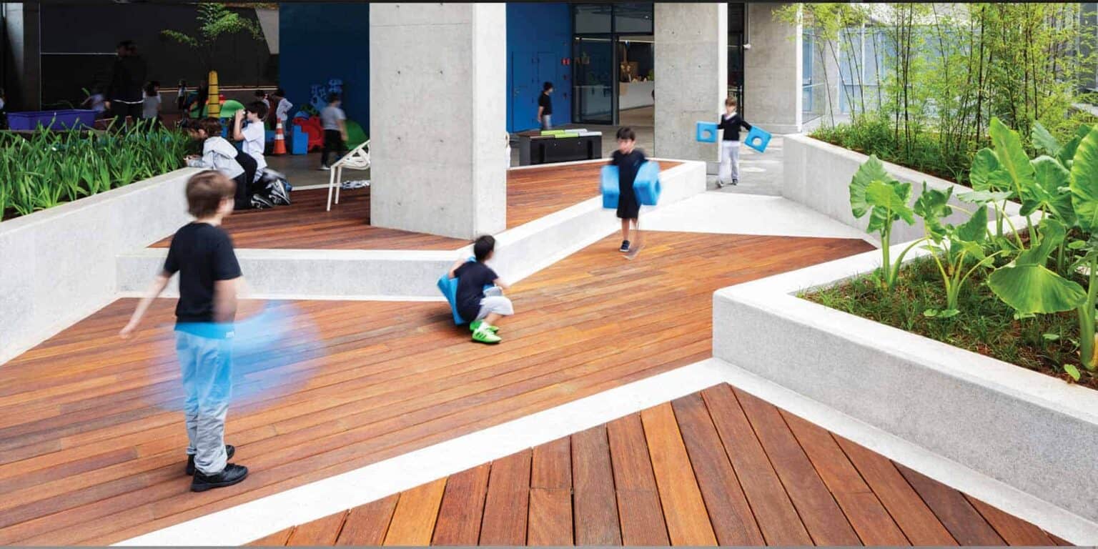 Bright modern school outdoor area with children playing on wooden decks and lush green plants, promoting active learning and well-being.
