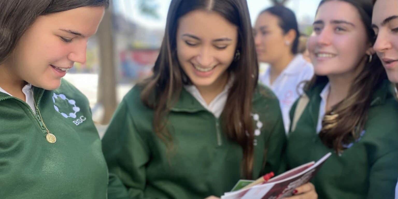 Smiling students in green school uniforms sharing a book outdoors at a World Schools event.
