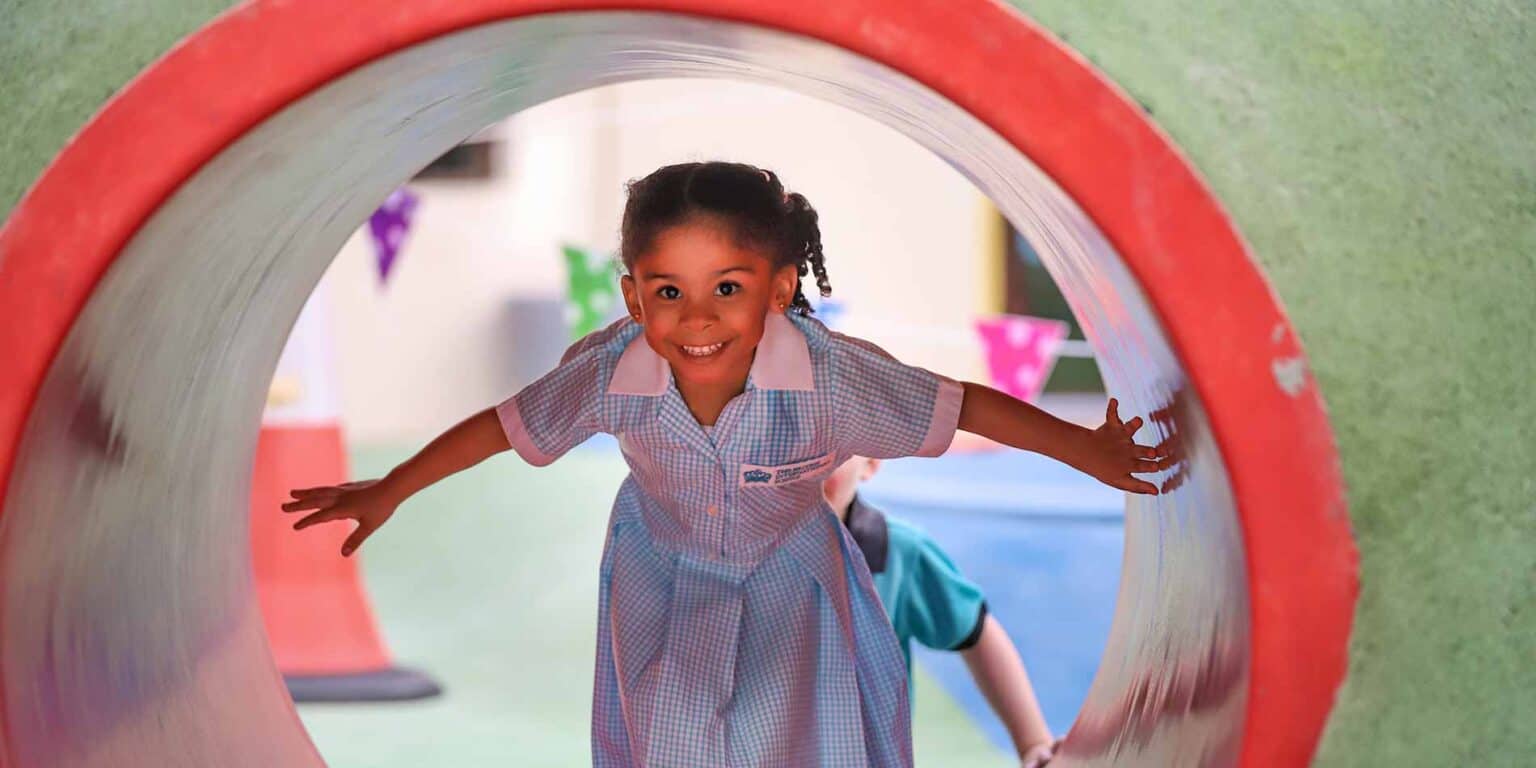 Niña de ojos brillantes con uniforme escolar gateando por el colorido túnel de juegos de una escuela internacional.