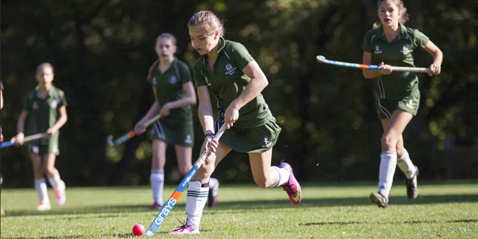 Girls playing field hockey outdoors at a school sports event under sunlight.