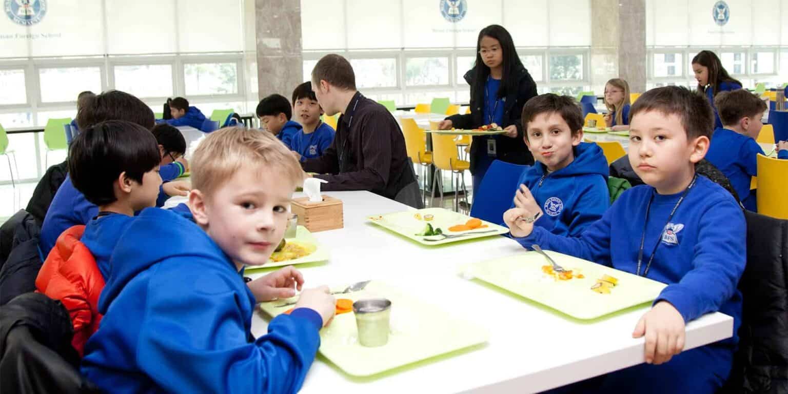 Ages children eating lunch in a modern school cafeteria with colorful chairs and large windows.