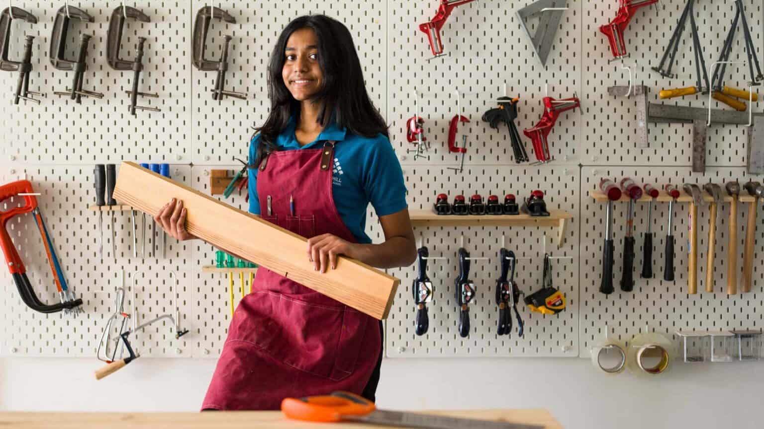 High school girl holding a wooden plank in a workshop with carpentry tools on wall.