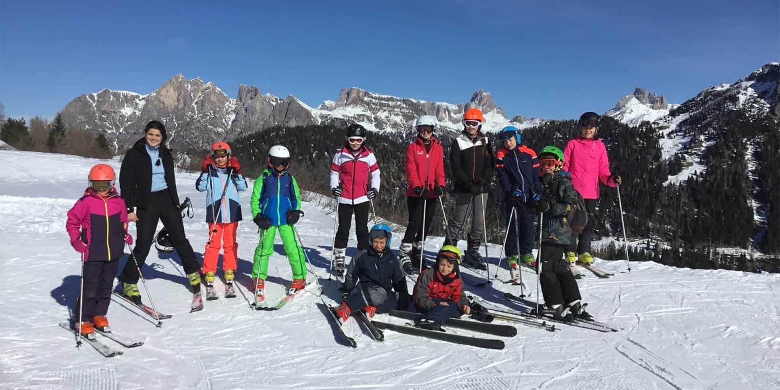 Alpine skiing school children in colorful gear on snowy mountain slope with skiers and scenic landscape.