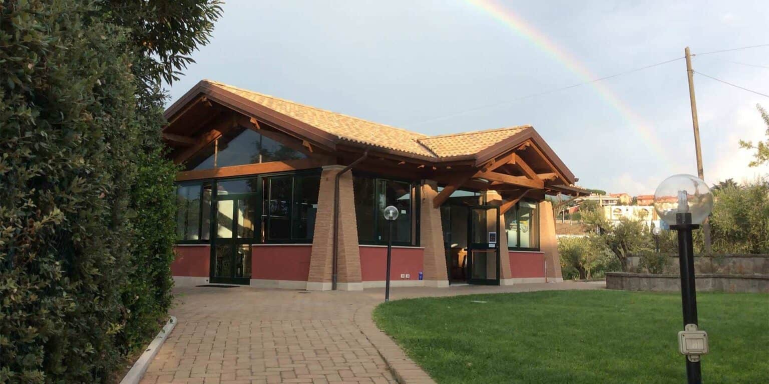 Modern school building with glass windows and wooden roof structure, set in lush greenery, under a rainbow sky.