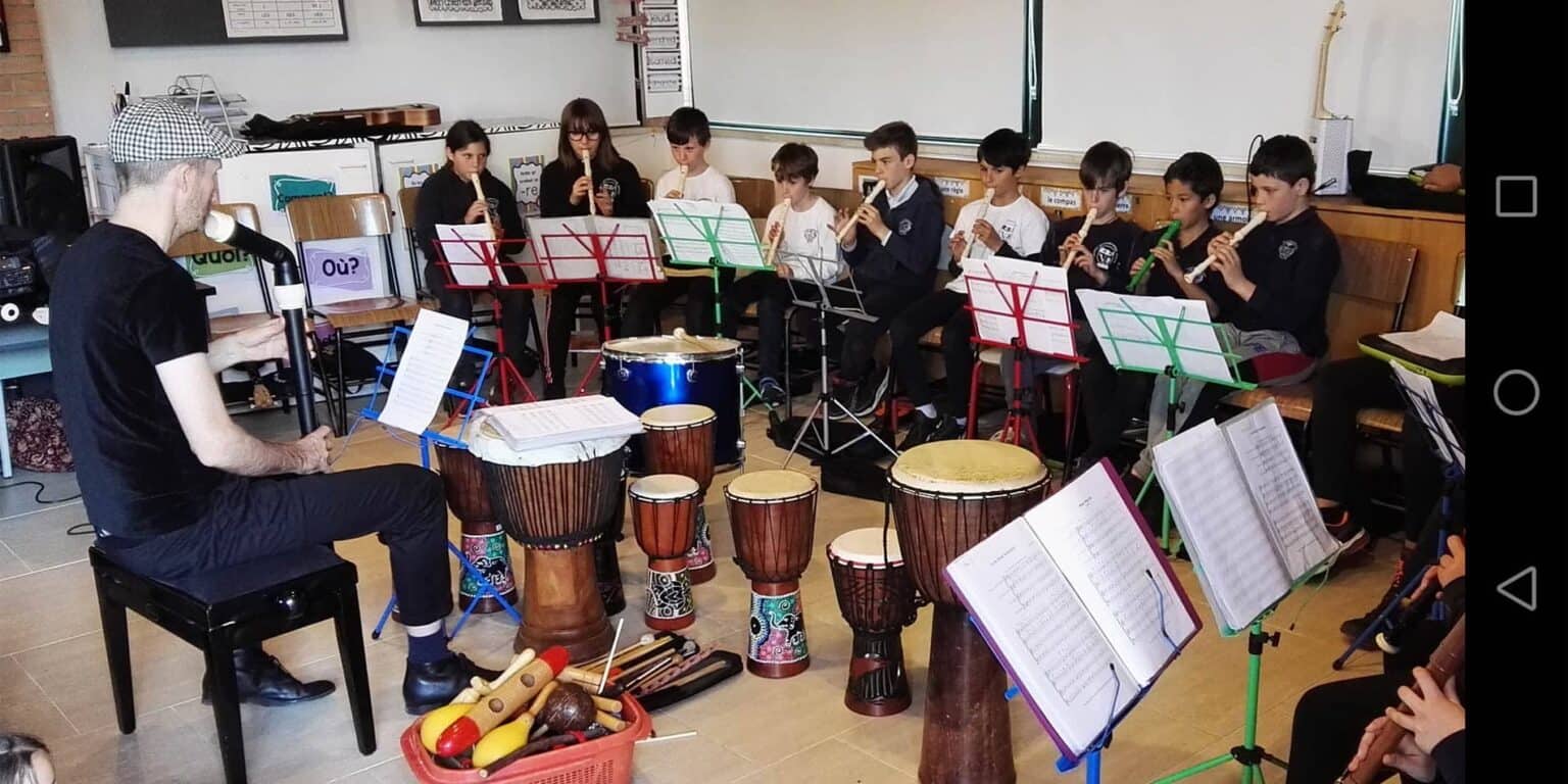 Children playing recorders in a school music class, with various drums and musical instruments nearby.