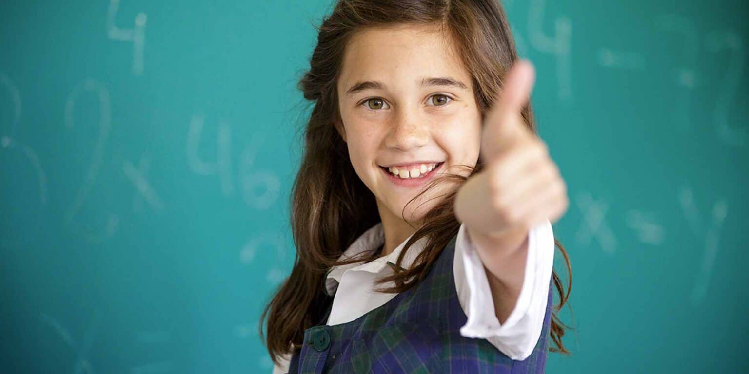 Bright schoolgirl giving a thumbs-up in classroom with chalkboard background, promoting global education and top-rated international schools.