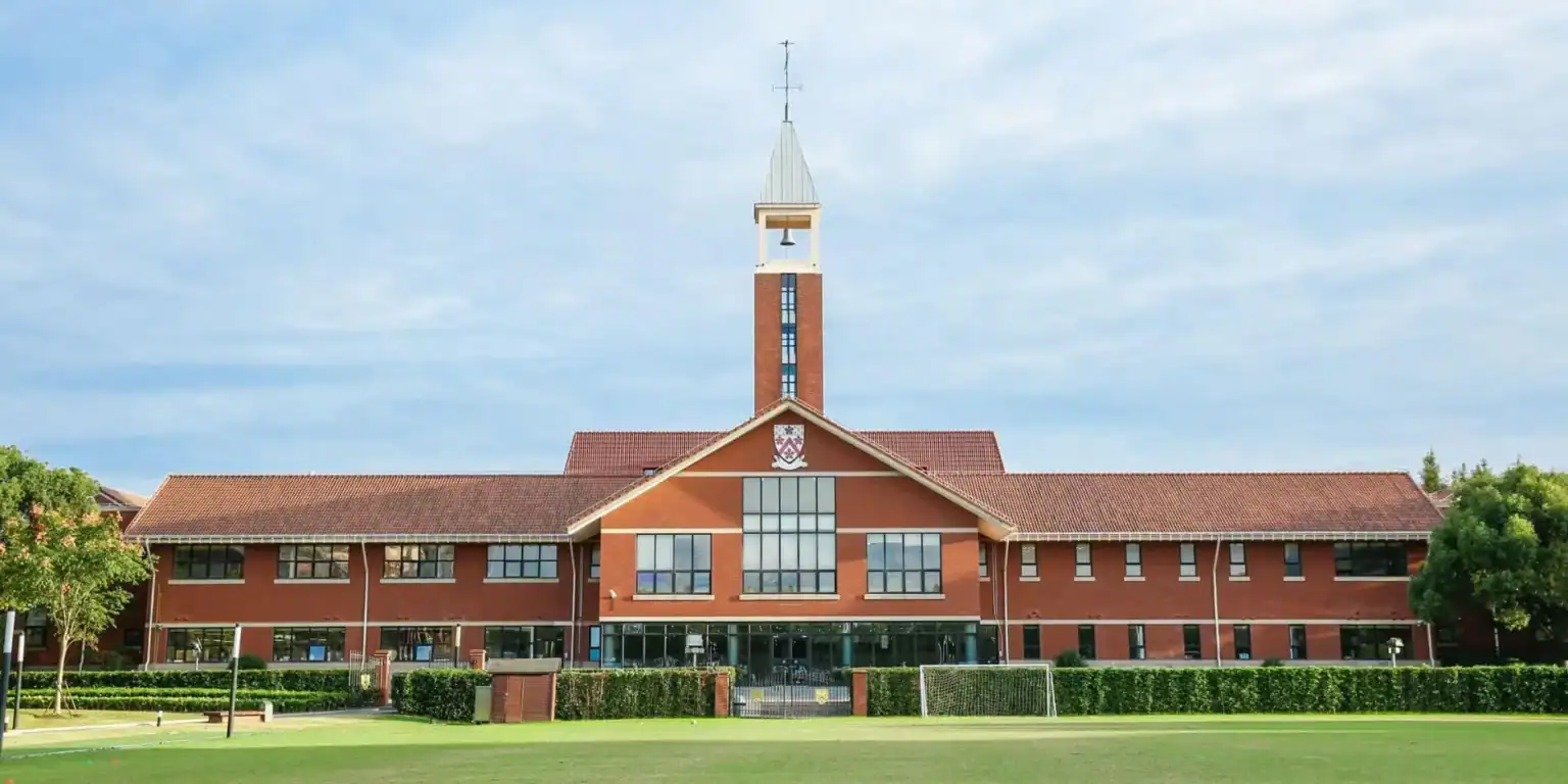 Modern school building with red brick facade, clock tower, and lush green field in front, part of a prestigious international school.