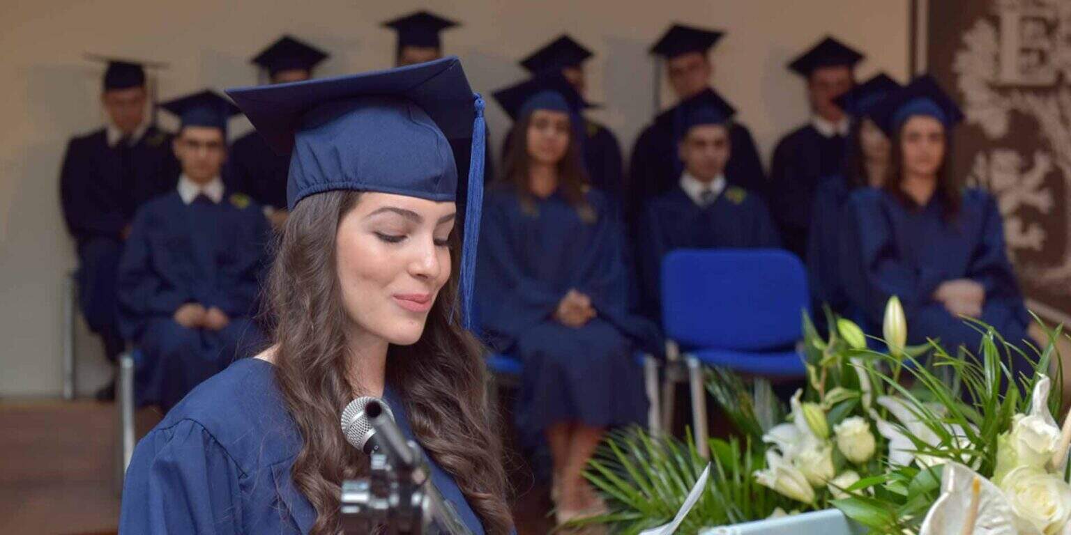 Graduate in cap and gown with closed eyes at graduation ceremony, diverse group in background.