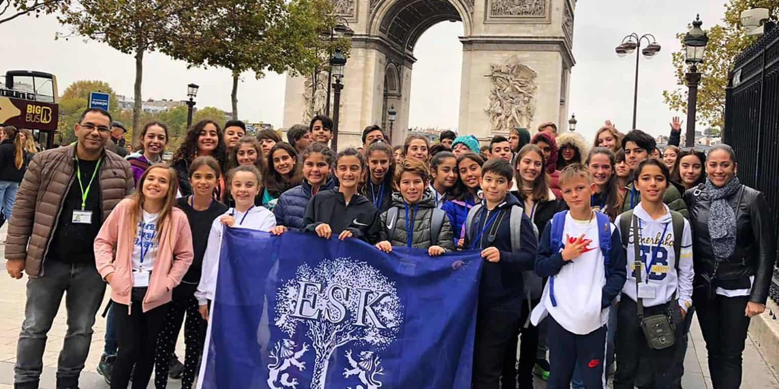 Bright group of diverse schoolchildren and teachers in Paris, holding a blue banner, at the Arc de Triomphe.