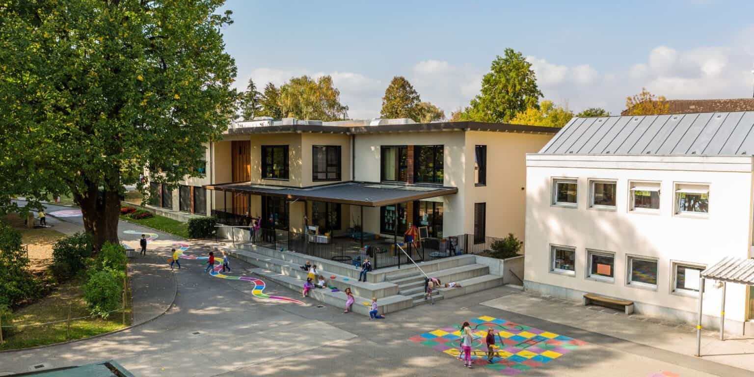 Bright modern school building surrounded by trees with children playing outside on colorful playgrounds.