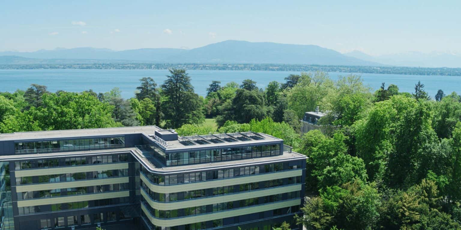 Modern school building surrounded by green trees with a lake and mountains in the background, showcasing scenic educational campus.