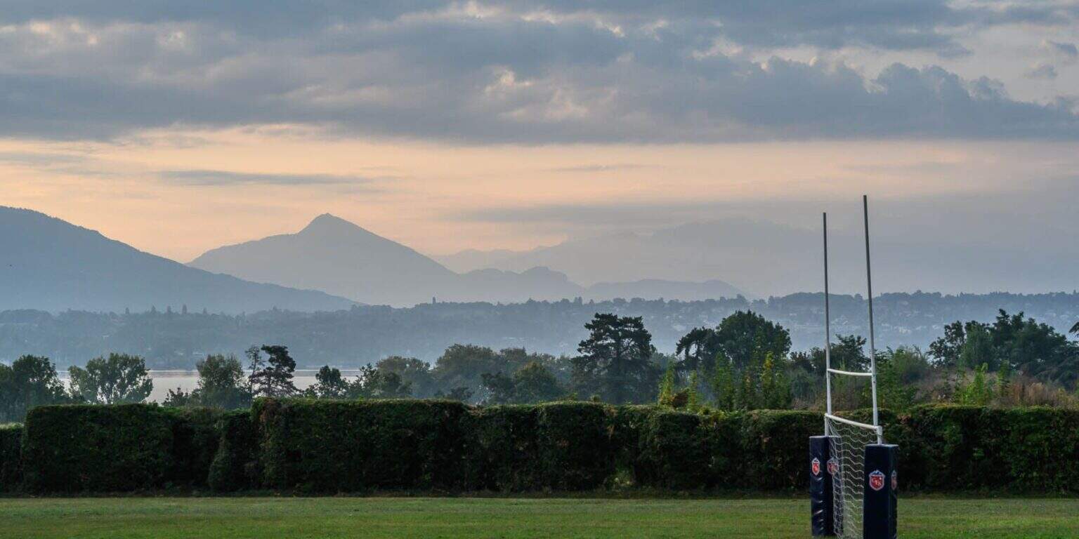 Mountains and rugby goalpost on a sports field with lush greenery and clouds in the sky.