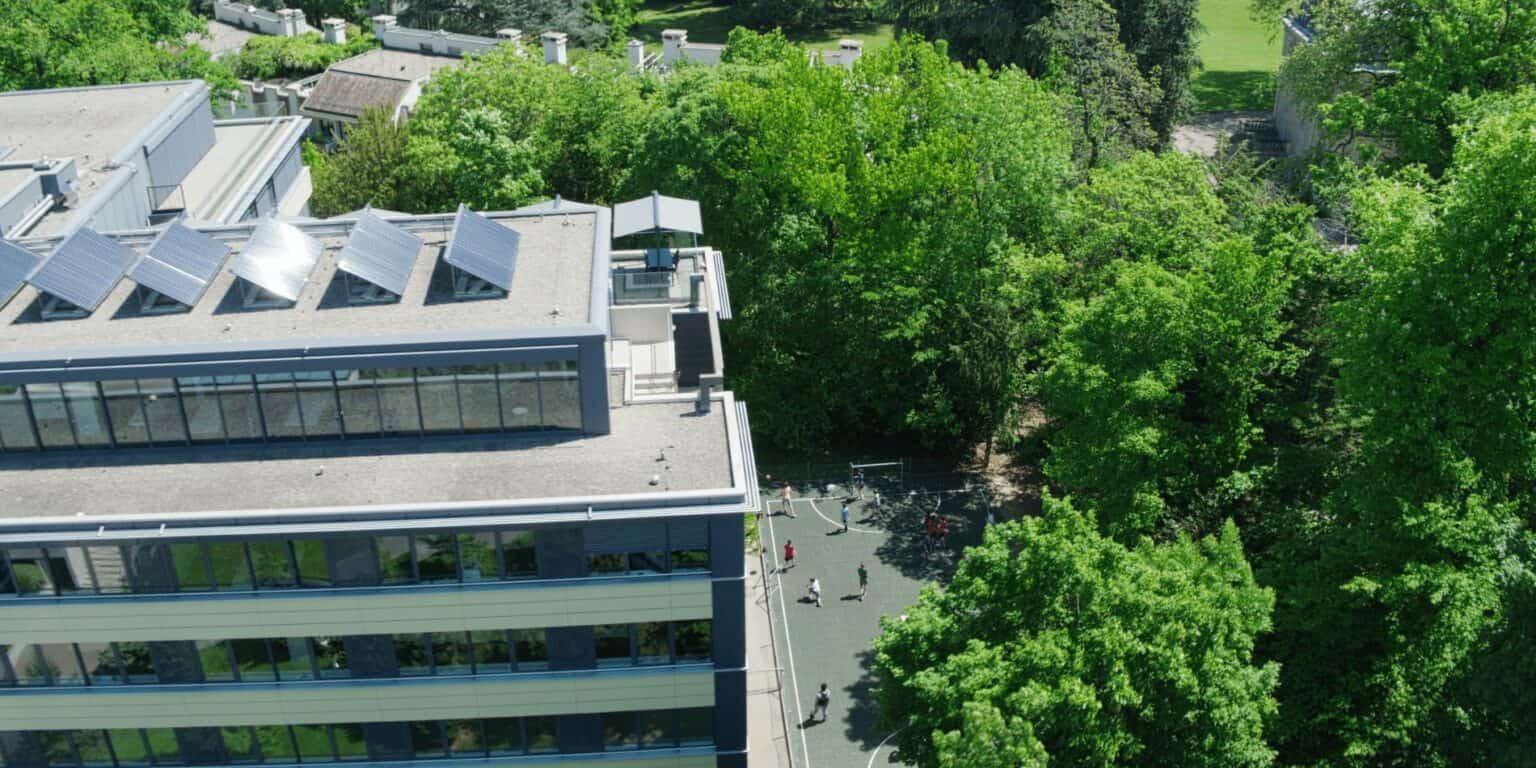 Aerial view of modern school building surrounded by green trees and students playing outside on a sunny day.
