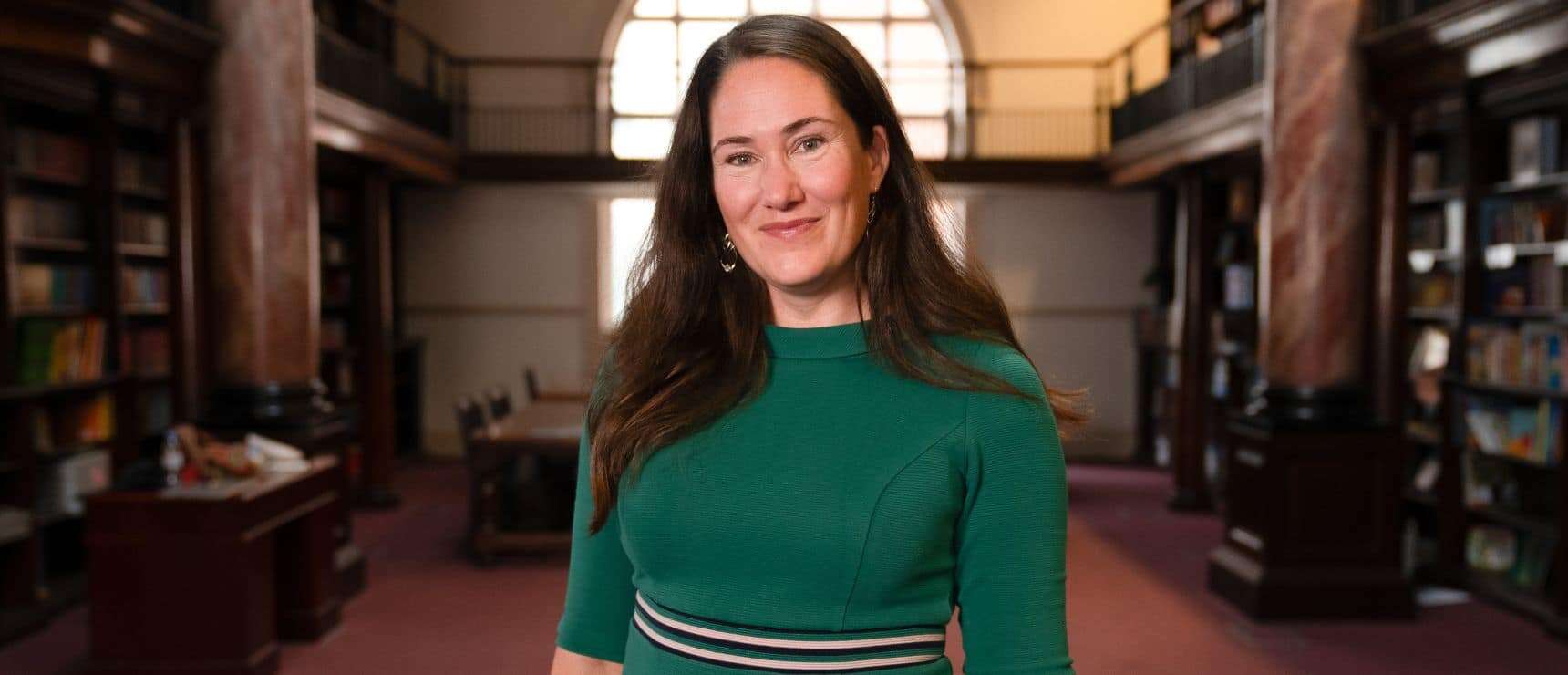 Bright smiling woman in a green dress standing in a historic library with tall bookshelves.