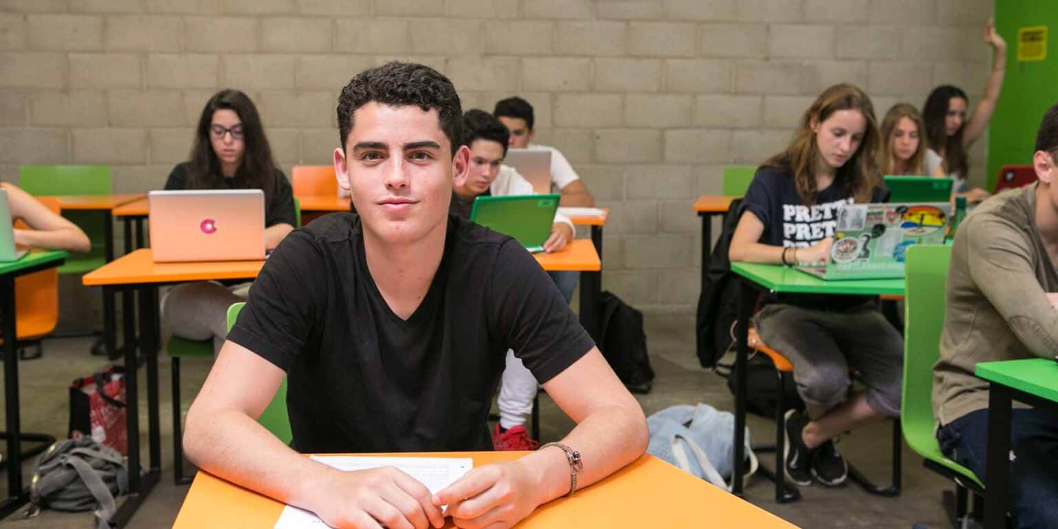 Young male student studying in a modern classroom with colorful desks and engaged peers.