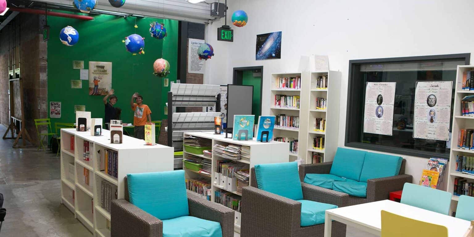 Colorful school library with bookshelves and students smiling and waving in front of a green wall.