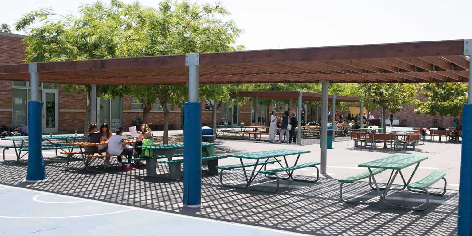 Students studying and socializing outdoors at a modern school campus with shaded picnic tables and greenery.