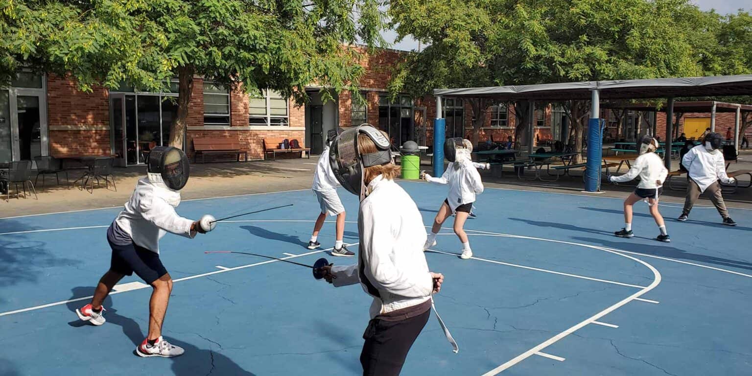 Children fencing practice outdoors at a school playground with blue court surface.