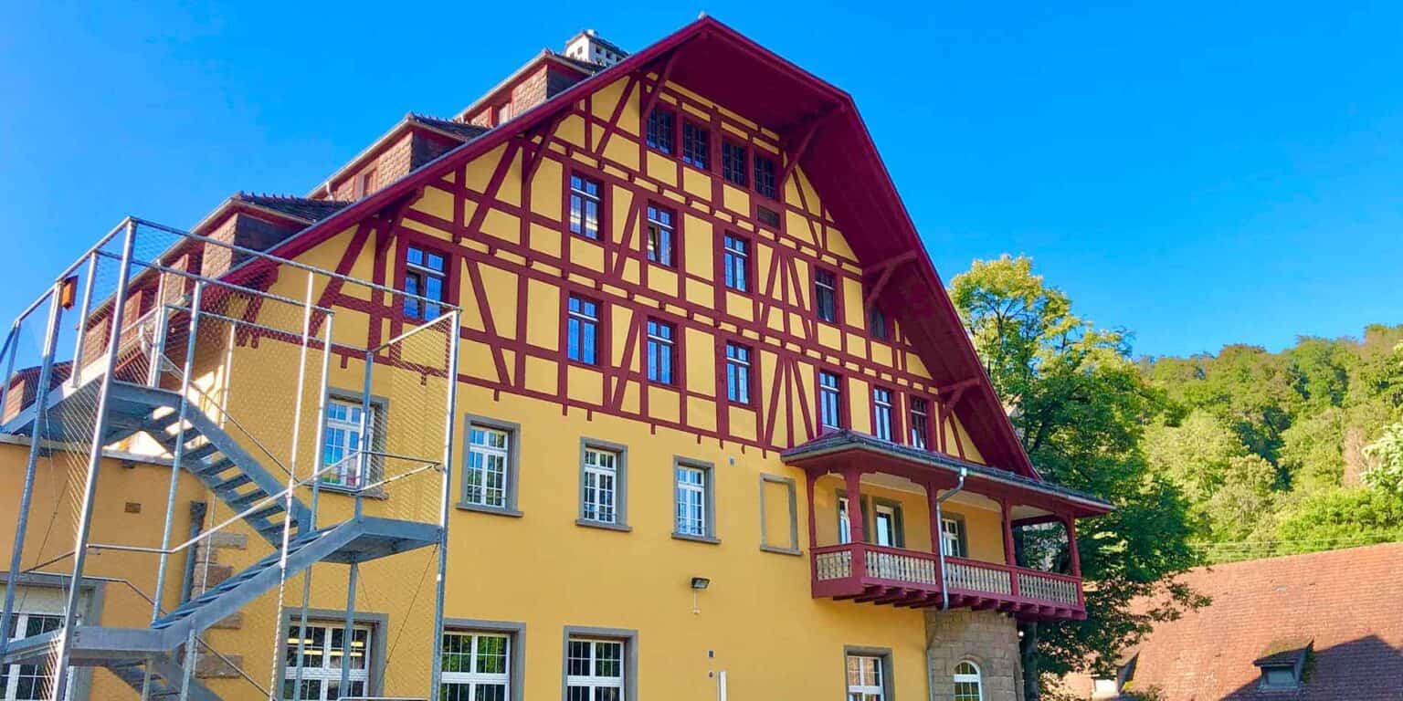 Colorful traditional European school building with bright yellow and red accents, surrounded by greenery.